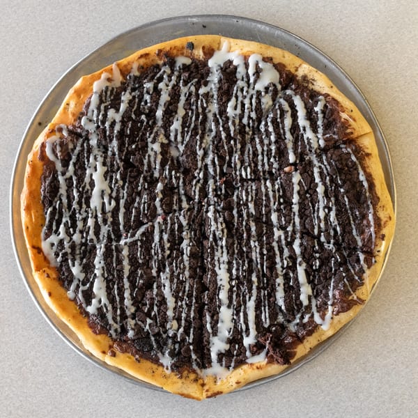 Dessert pizza with chocolate cookie crumbles and white icing drizzle on a metal pan.