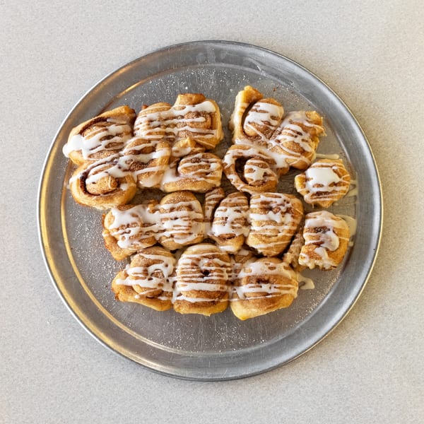 A silver platter filled with numerous cinnamon rolls drizzled with white icing, on a light gray surface.