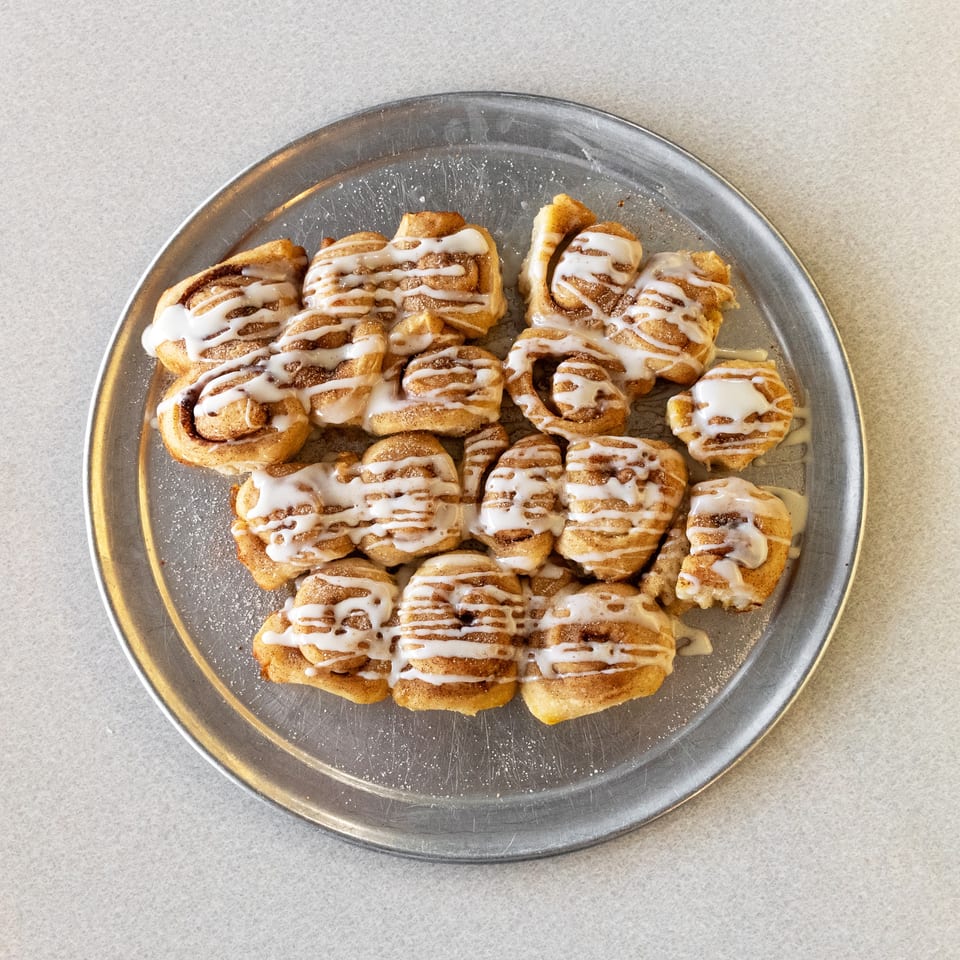 A silver platter filled with numerous cinnamon rolls drizzled with white icing, on a light gray surface.